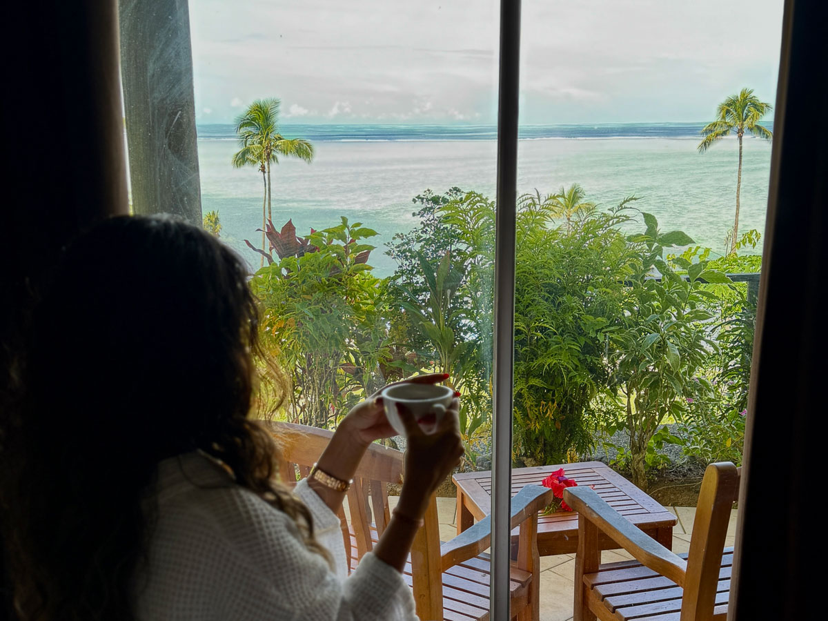 Woman enjoys cup of tea looking out window of Deluxe Seaview Bure at Crusoe's Retreat, Fiji